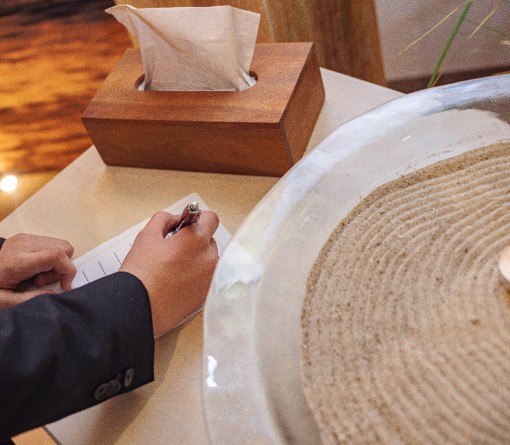 Schreibende Männerhand. Auf dem Tisch zusätzlich Kleenex-Box aus Holz und Glasschale mit Dekosand.