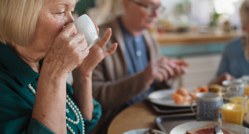 Ältere Dame am Tisch sitzend von der Seite mit Kaffeetasse am Mund, im Hintergrund weitere Personen unscharf.