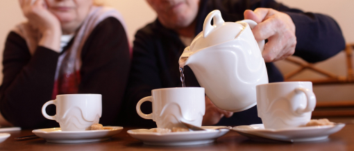 Drei weiße Tassen auf Tisch. Älteres Ehepaar unscharf im Hintergrund. Er schenkt Tee ein.