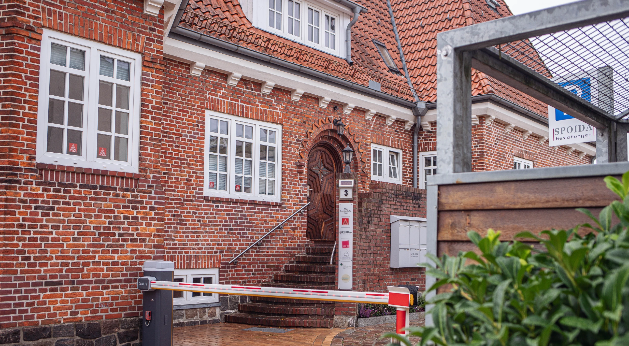Treppe Backsteingebäude mit Firmenschildern. Schranke und Busch im Vordergrund.