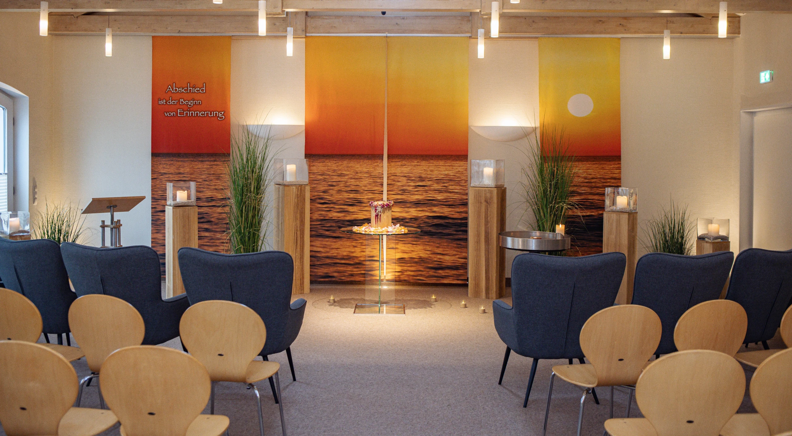 Trauerhalle, links rechts Stühle, vorne Sessel. Kerzenstehlen, Urne mittig mit Teelichtern, Hintergrund Sonnenuntergang.
