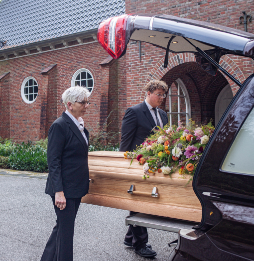 : Zwei Mitarbeiter, Mann und Frau, laden hellen Sarg mit Blumengesteck in Bestattungswagen. Hintergrund Backsteinkirche.