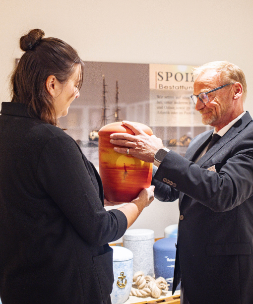 : George Spoida übergibt Urne mit Sonnenuntergang am Meer in Orange an Mitarbeiterin.