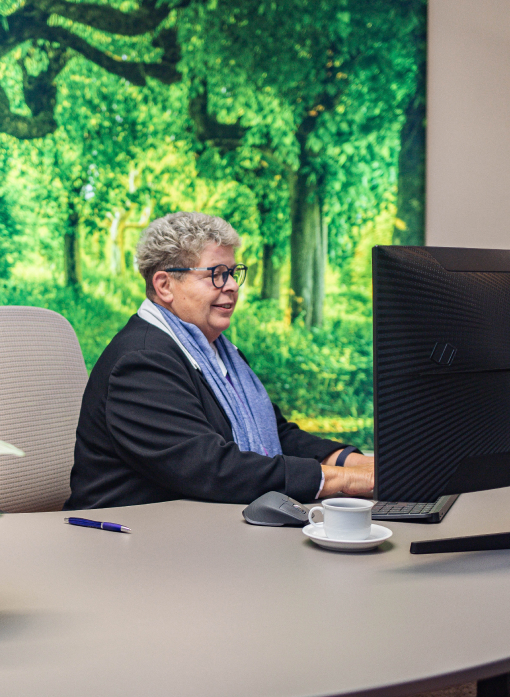 Mitarbeiterin am Computer von der Seite, vor Tastatur sitzend, Stift und Kaffeetasse, hinten Waldbild.
